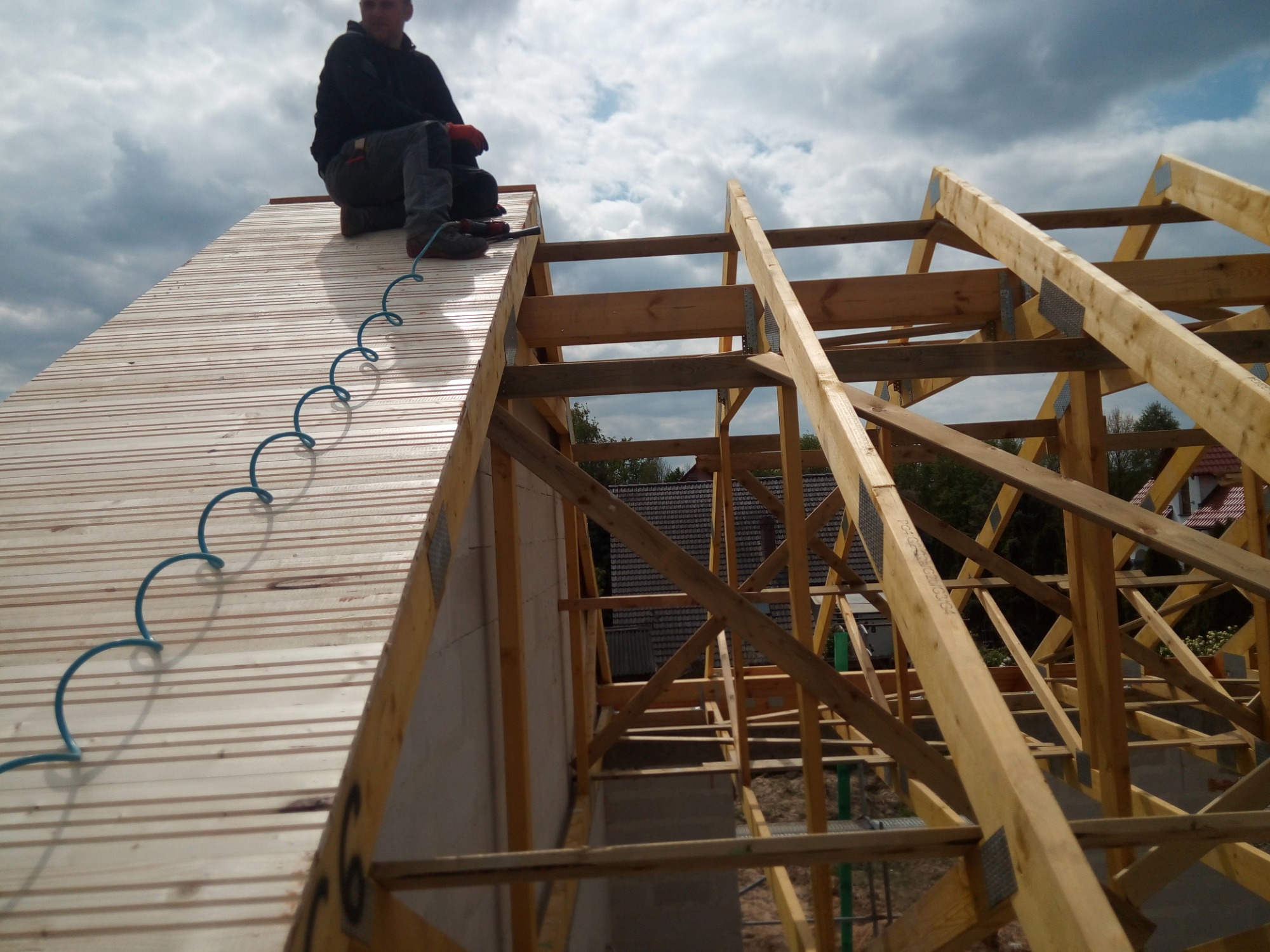 Pracownik siedzi na drewnianym poszyciu dachu, obok widoczna konstrukcja więźby dachowej i zwisający niebieski przewód pneumatyczny.
