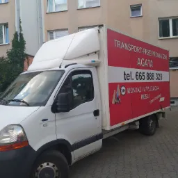 Biały samochód dostawczy z czerwonym nadrukiem reklamowym firmy transportowej 'Agata' z numerem telefonu, stojący na chodniku przed budynkiem mieszkalnym.