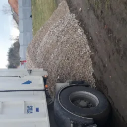 Ciężarówka STAS wyładowuje żwir na ziemię w pobliżu ogrodzenia z betonowych płyt, widoczne ślady opon w błocie.