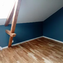 Poddasze po remoncie: ciemnoniebieskie ściany, drewniane belki konstrukcyjne i jasny parkiet, widok zza uchylonych drzwi.