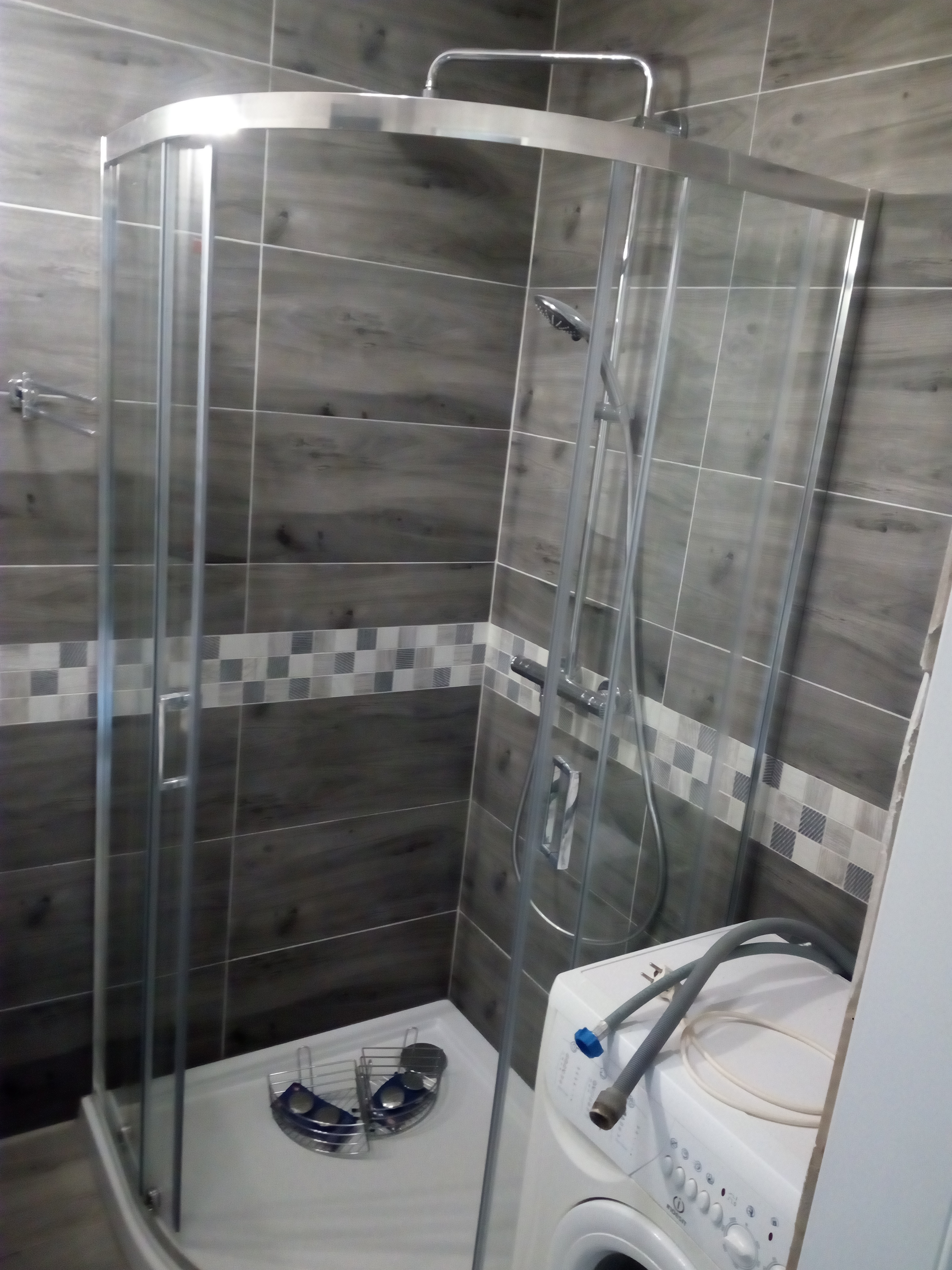 Narożna kabina prysznicowa z chromowanymi profilami i deszczownicą, zamontowana w łazience z szarymi płytkami imitującymi drewno i dekoracyjnym pasem mozaiki, obok pralka z podłączonymi wężami.