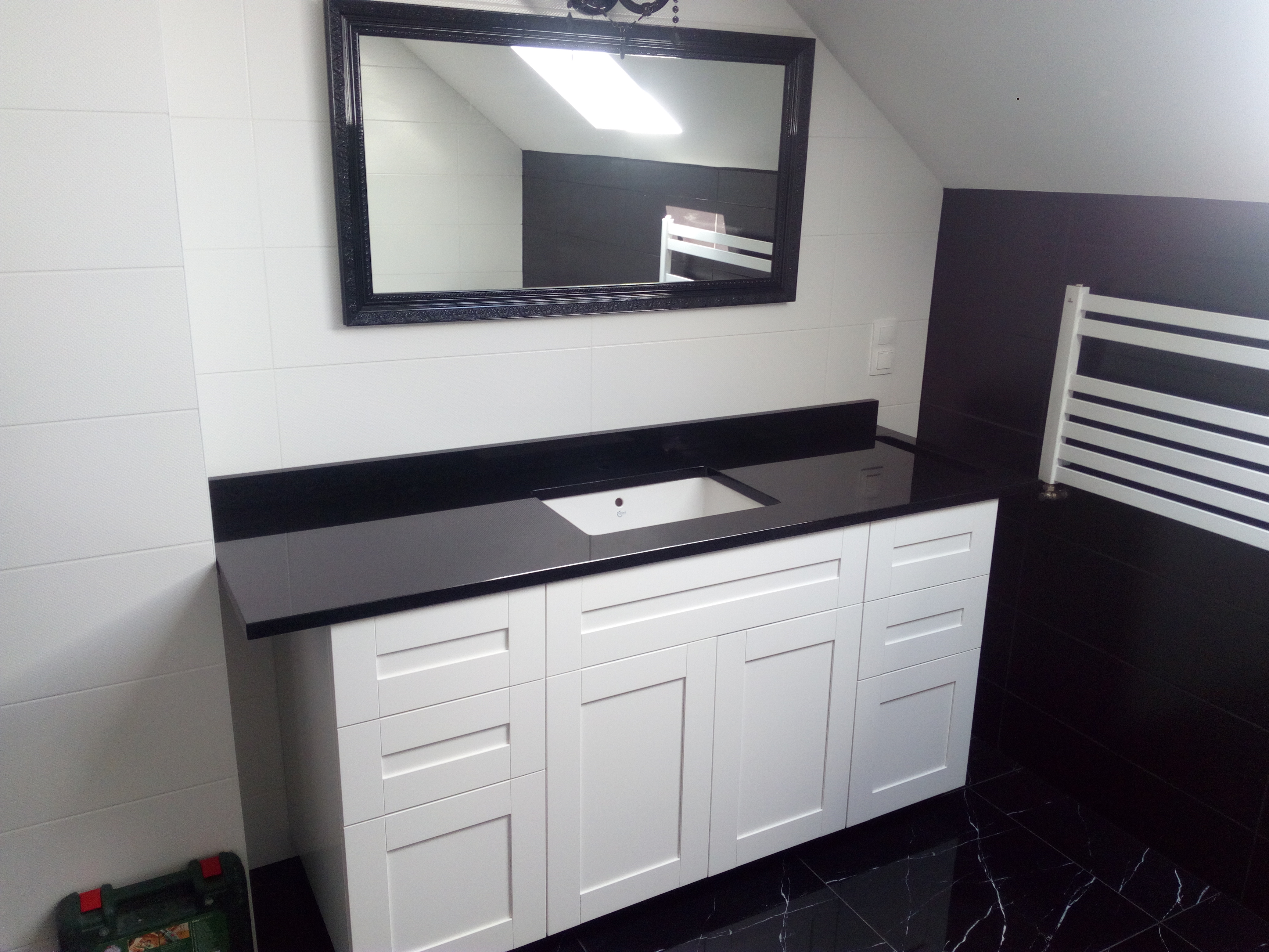 Biała szafka łazienkowa z czarnym blatem i umywalką, duże czarne lustro w ozdobnej ramie, czarno-białe płytki na ścianach i podłodze, grzejnik drabinkowy.