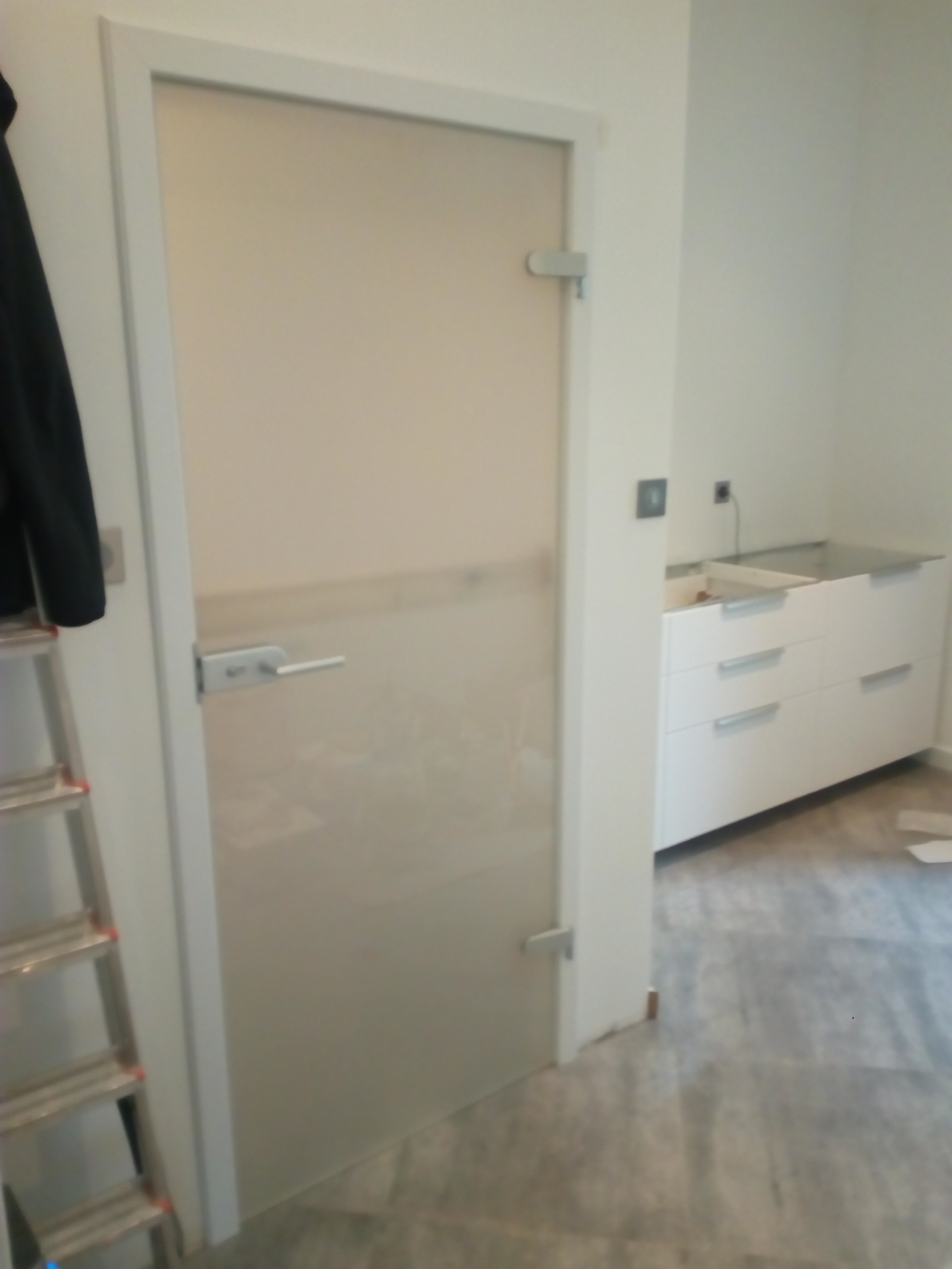 Szklane drzwi wewnętrzne z białą ościeżnicą, widoczne białe szafki z szufladami oraz drabina malarska w tle.