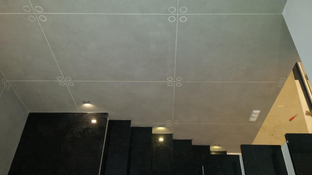 Szara, gładka powierzchnia sufitu podzielona białymi liniami z dekoracyjnymi rozetami w punktach przecięcia, oświetlona małymi, kwadratowymi lampkami; poniżej widoczne czarne, błyszczące stopnie...