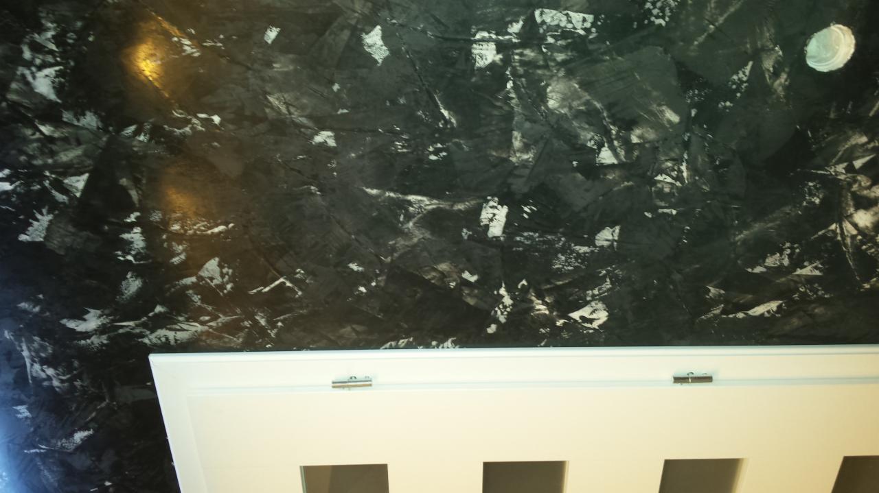 Czarno-biała, abstrakcyjna faktura na ścianie z widocznym fragmentem białej ramy drzwiowej z metalowymi zawiasami.