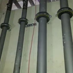 Cztery pionowe rury wentylacyjne z czarnymi kolankami i ż&oacute;łtymi uszczelkami, zamontowane przy szarej ścianie w surowym wnętrzu z widocznymi kablami pod sufitem.