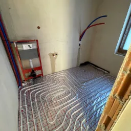 Instalacja rur grzewczych na folii izolacyjnej w surowym wnętrzu łazienki przed wylewką, widoczny stelaż podtynkowy WC i wystające rury instalacyjne.