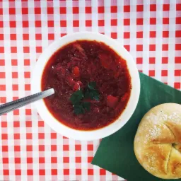 Czerwony barszcz ukraiński w białej misce, podany z bułką na zielonej serwetce i łyżką, na czerwono-białym obrusie w kratę.