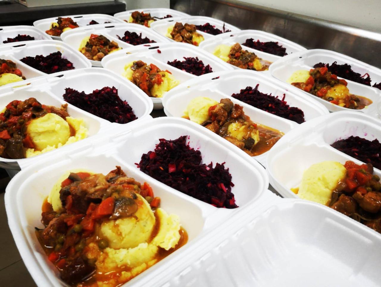 Rzędy styropianowych pojemników z gotowymi posiłkami: puree ziemniaczane z gulaszem warzywno-mięsnym i buraczkami na białym stole.