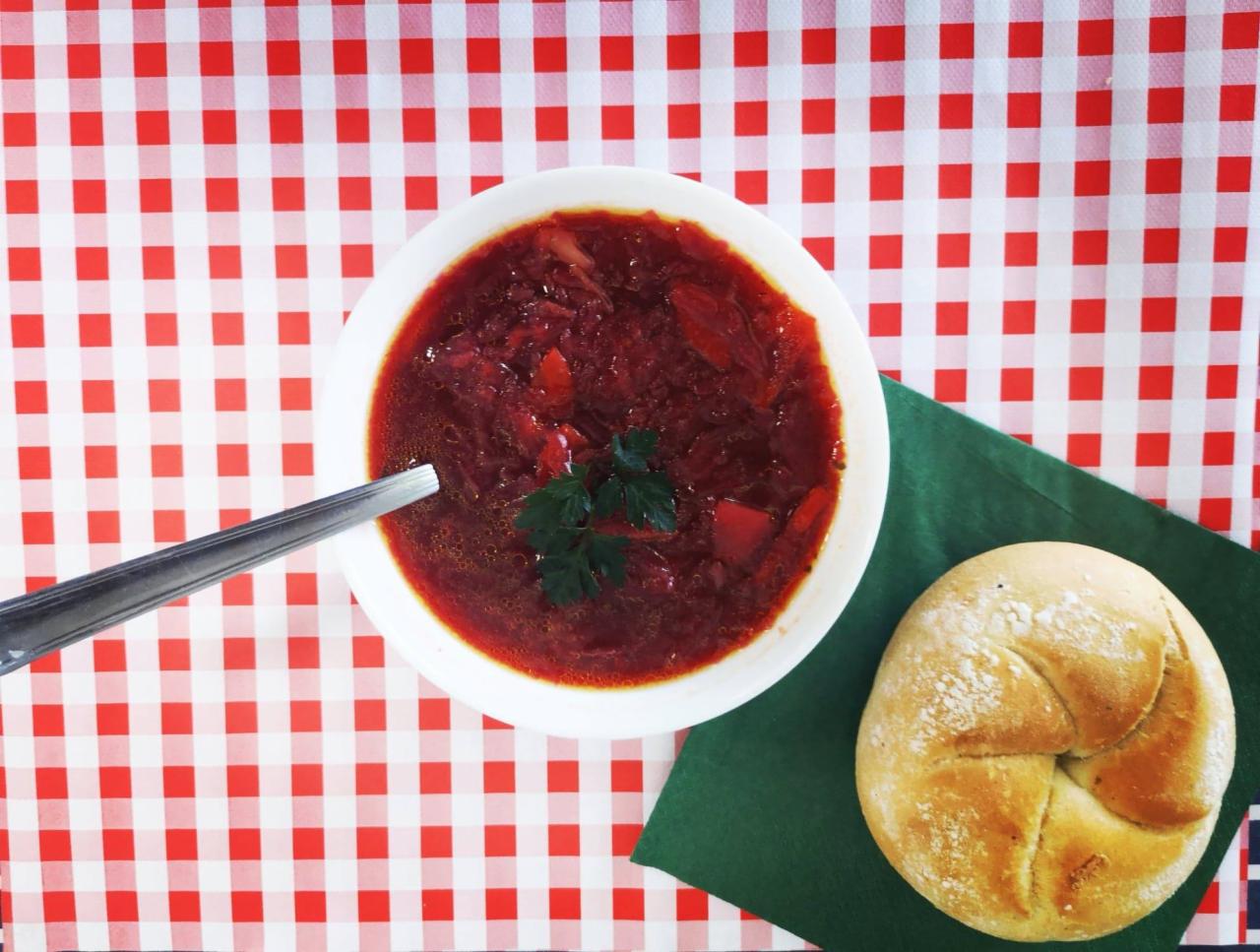 Czerwony barszcz ukraiński w białej misce, podany z bułką na zielonej serwetce i łyżką, na czerwono-białym obrusie w kratę.