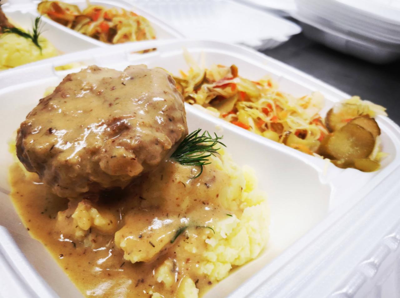 Kotlet mielony w sosie, ziemniaki puree i surówka z kapusty w białym, plastikowym pojemniku na wynos, udekorowane gałązką koperku.