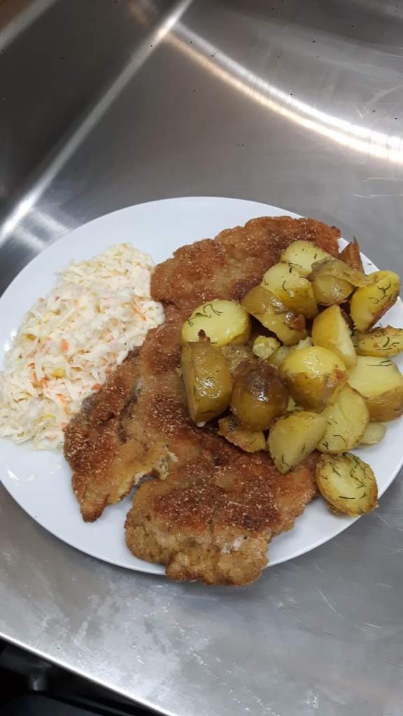 Dwa panierowane kotlety schabowe z ziemniakami posypanymi koperkiem i surówką z białej kapusty na białym talerzu, ustawione na metalowym blacie.