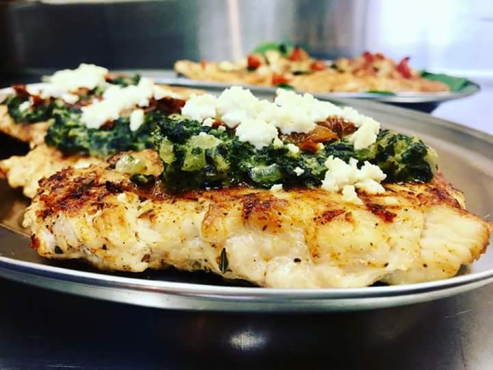 Grilowana ryba z dodatkiem szpinaku, suszonych pomidorów i sera feta na metalowym talerzu, w tle inne danie.