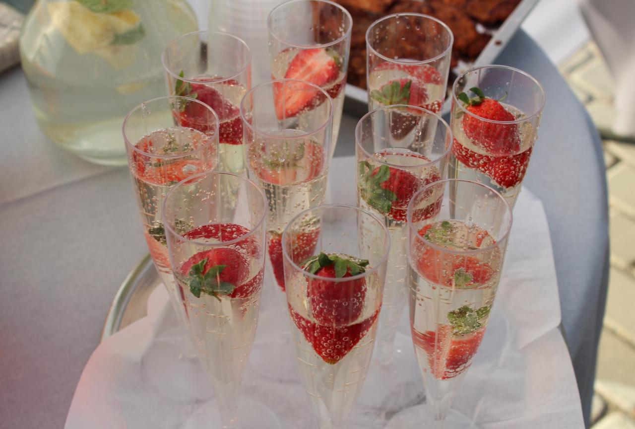 Ustawione na srebrnej tacy kieliszki z szampanem i truskawkami, z widocznym dzbankiem z napojem i przekąskami w tle.