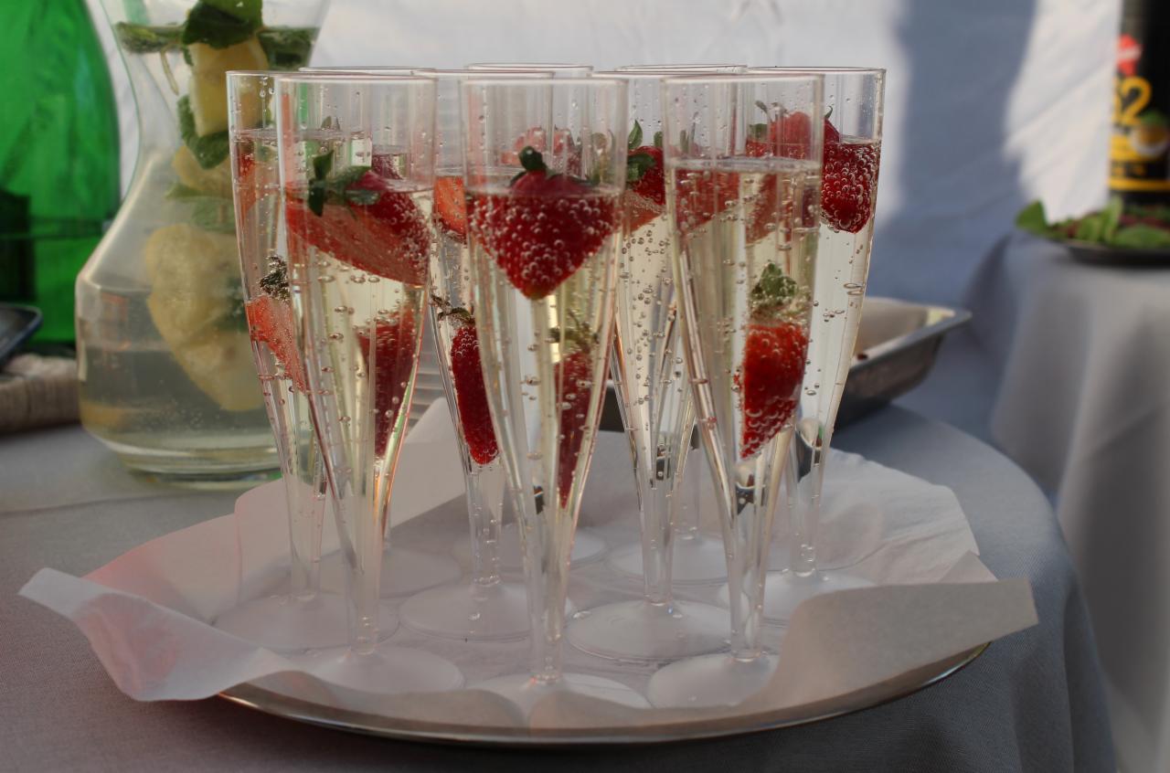 Szereg kieliszków do szampana wypełnionych musującym napojem z pływającymi kawałkami truskawek, ustawionych na srebrnej tacy z serwetkami, w tle dzbanek z lemoniadą.