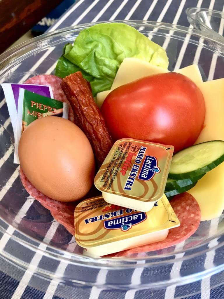 Pojemnik z przekąskami: jajko na twardo, pomidor, ser żółty w plastrach, plasterki salami, ogórek, sałata, kiełbaska myśliwska i dwie porcje masła w opakowaniach jednostkowych, widoczne przyprawy...