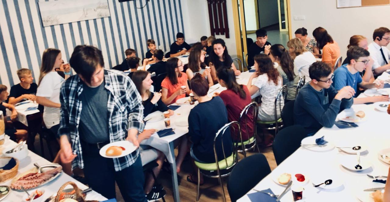 Grupa młodych ludzi siedząca przy stołach w sali, jedząca posiłek; jeden z mężczyzn w koszuli w kratę podaje talerz z jedzeniem; w tle ściana w biało-niebieskie pasy.