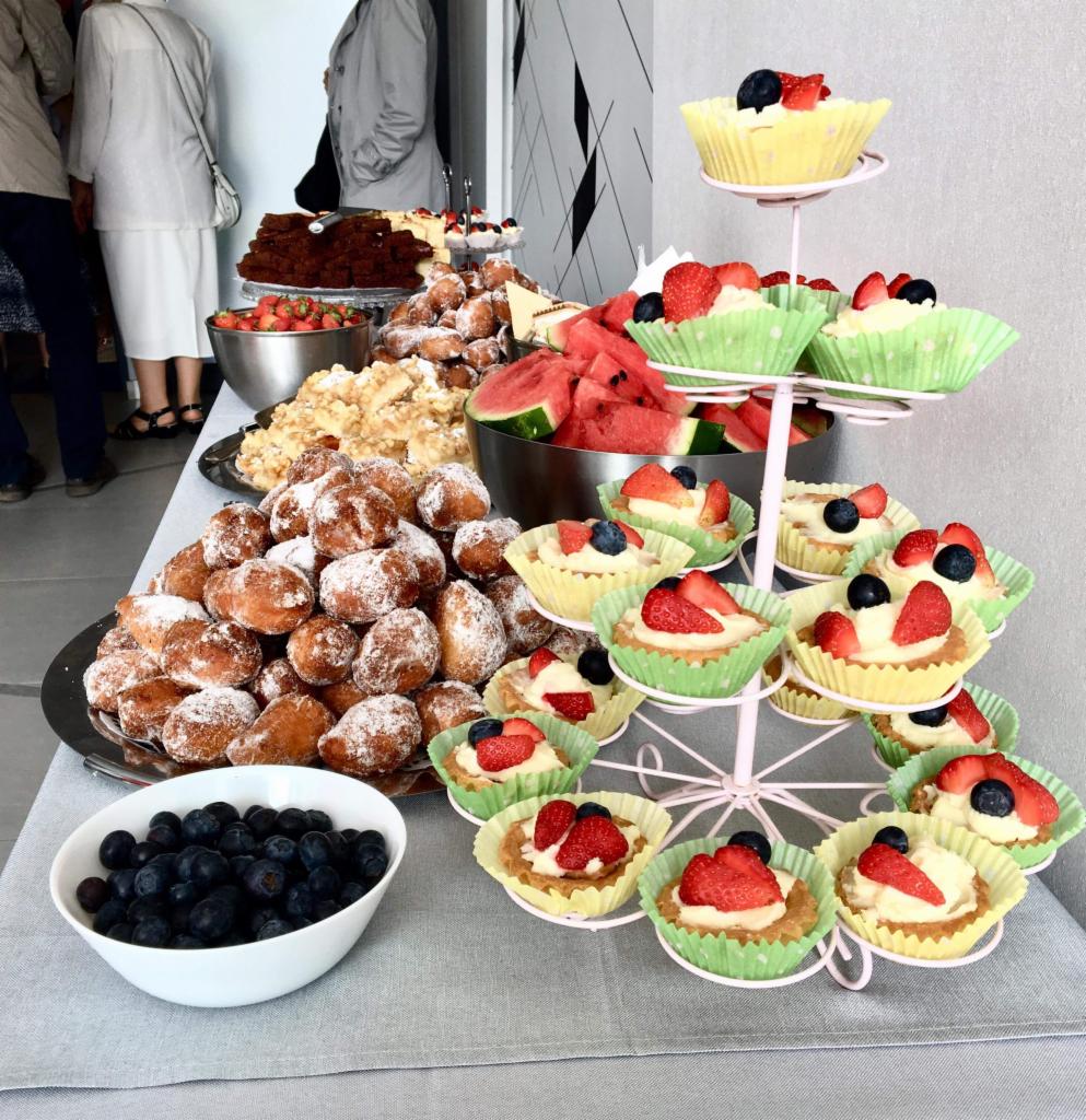 Stół zastawiony różnorodnymi deserami: pączki posypane cukrem pudrem, tartaletki z kremem i owocami (truskawki, borówki), arbuz pokrojony w trójkąty, ciasto czekoladowe, miseczka z borówkami...