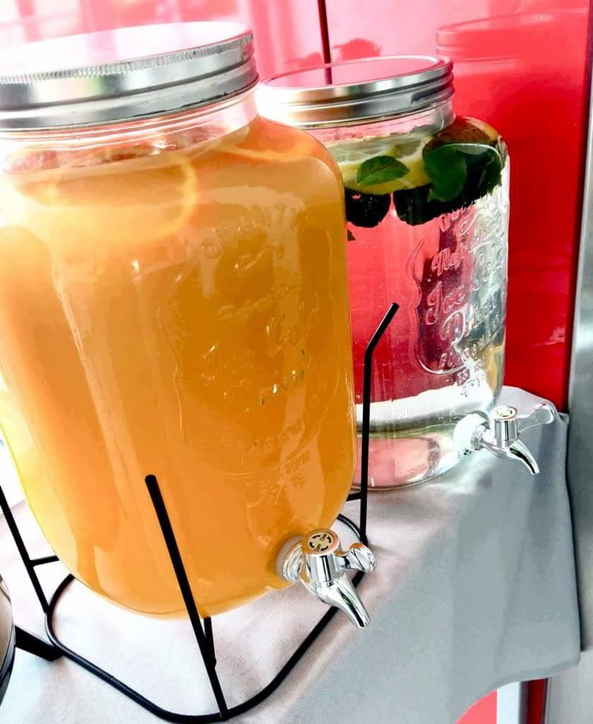 Dwa szklane słoje z kranikami, jeden z pomarańczowym napojem z plastrami pomarańczy, drugi z wodą, miętą i ciemnymi owocami, ustawione na stole z białym obrusem.