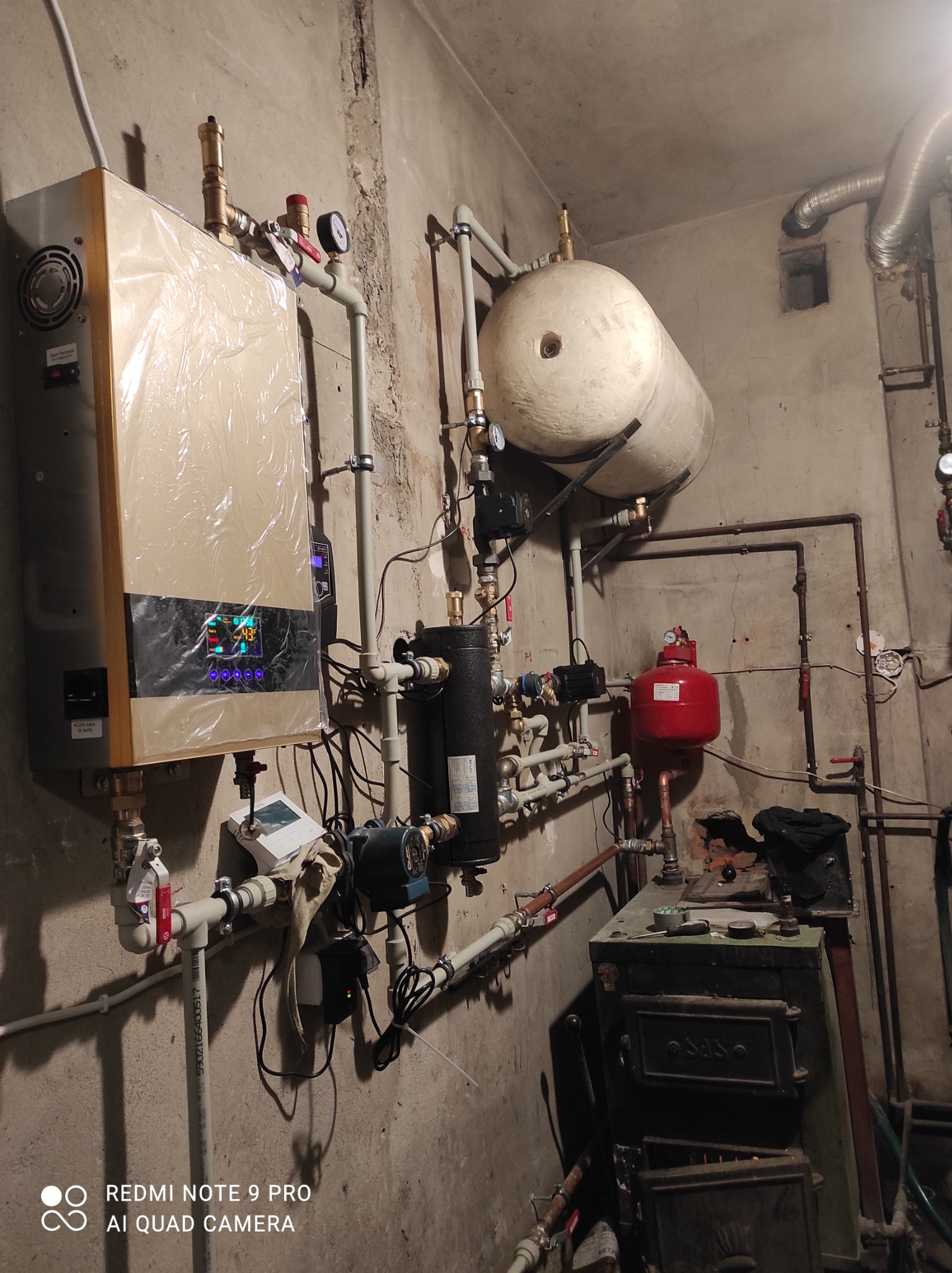 Nowoczesny piec grzewczy w folii ochronnej, podłączony do instalacji z rur miedzianych i plastikowych, zbiornik wyrównawczy nad piecem węglowym w surowym pomieszczeniu kotłowni.