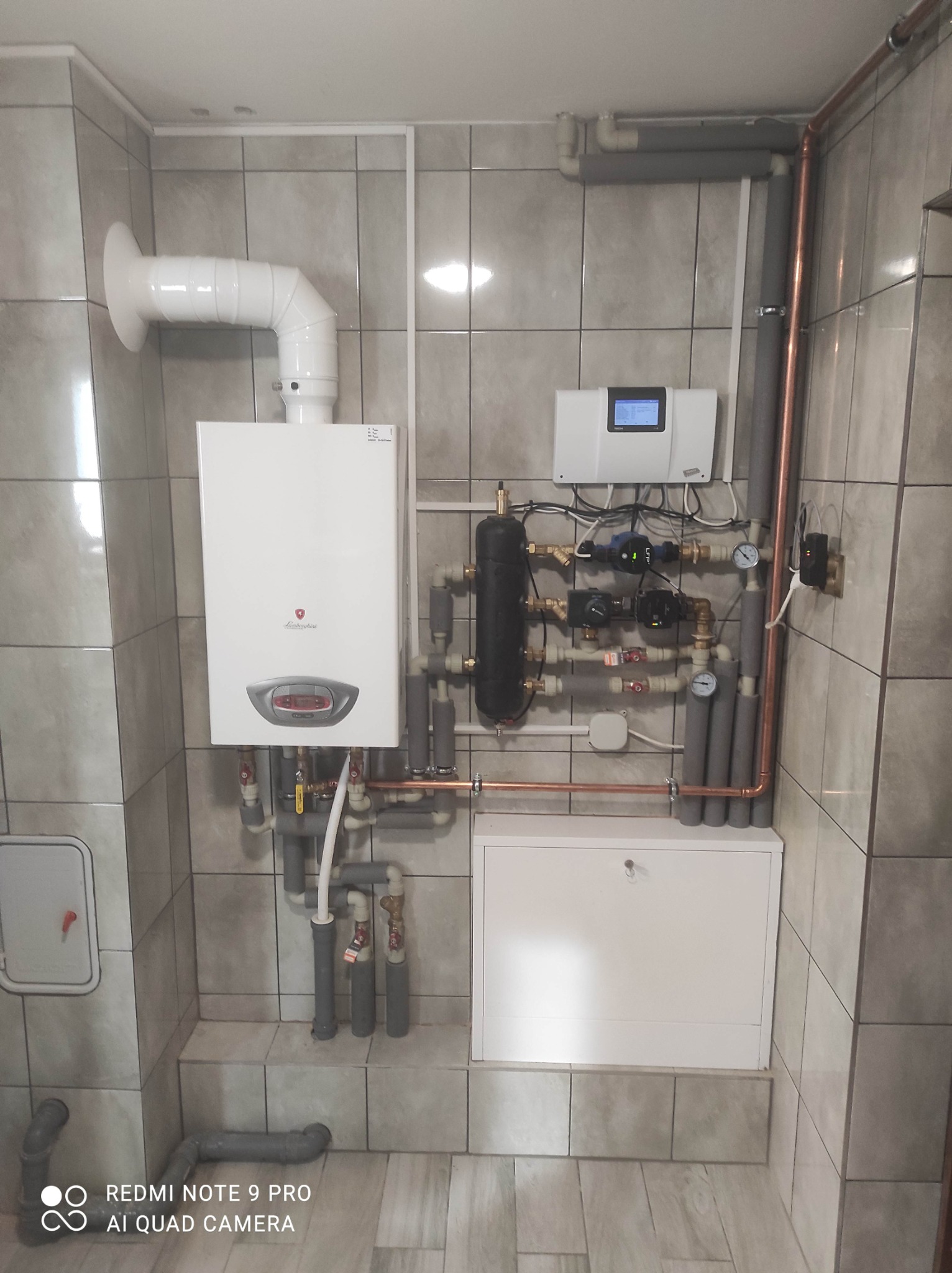 Nowoczesny piec gazowy z podłączeniami, rurami miedzianymi i białym panelem sterowania zamontowany na szarych płytkach w łazience.