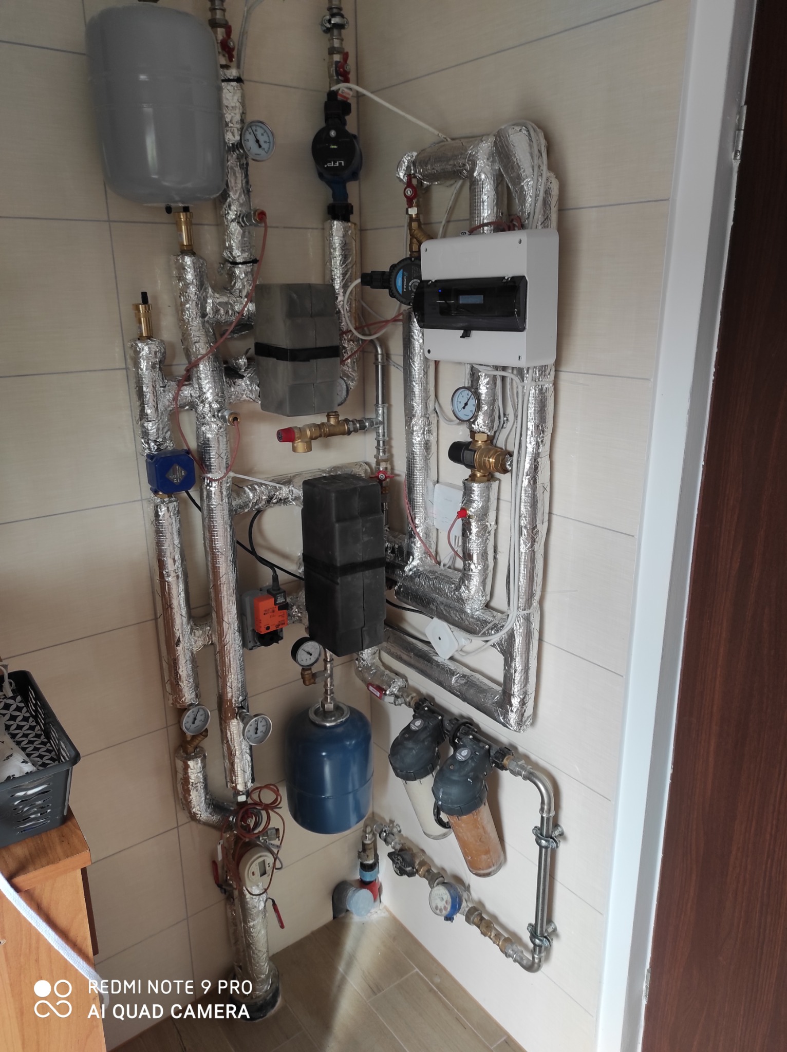 Złożona instalacja z rurami zaizolowanymi folią aluminiową, licznymi zaworami, manometrami, zbiornikami wyrównawczymi i filtrami wody, zamontowana na ścianie z płytek ceramicznych.