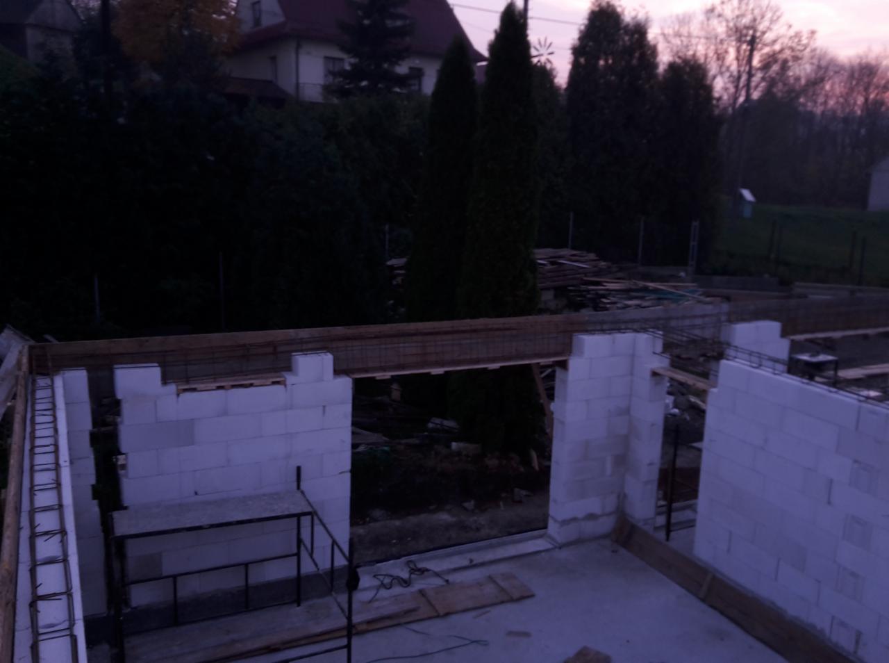 Wznoszenie ścian fundamentowych z bloczków betonowych na budowie domu jednorodzinnego, widoczne zbrojenie i szalunki, w tle drzewa i budynki sąsiedzkie o zmierzchu.