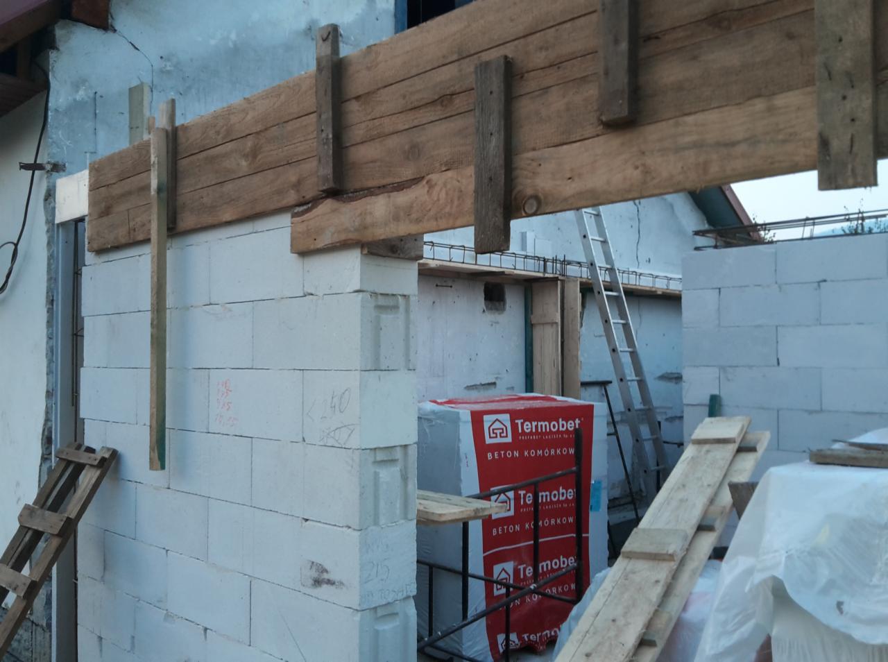 Wznoszenie ścian z betonu komórkowego, widoczne drewniane szalunki nad otworem, paleta z bloczkami Termobet i drabina aluminiowa w tle.