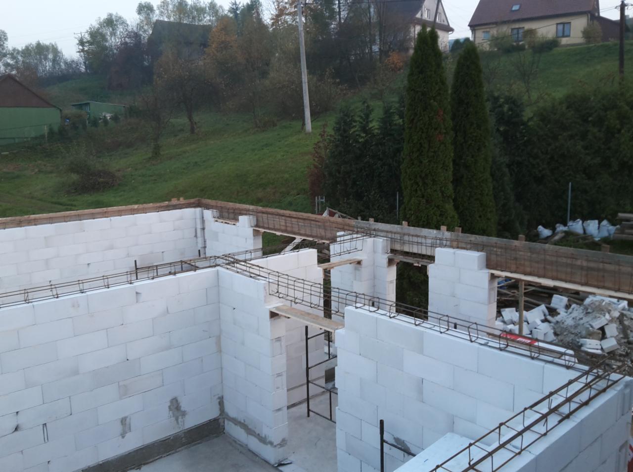 Wznoszenie ścian domu z białych bloczków, widoczne zbrojenia i drewniane elementy szalunku, w tle zielone wzgórze i zabudowa mieszkalna.