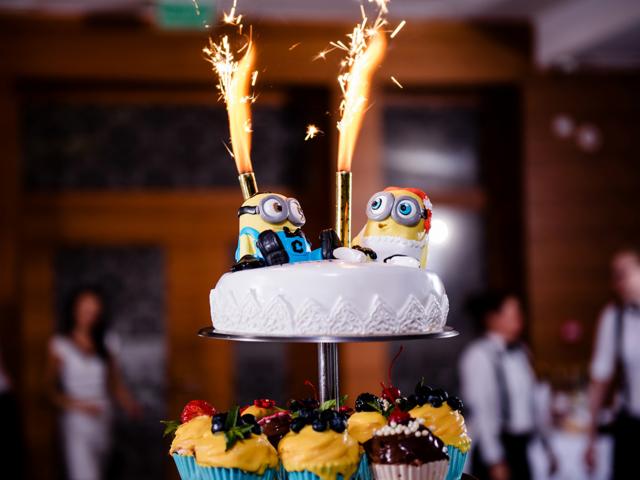 Dwupoziomowy tort weselny z dekoracją Minionków i fontannami iskier, na paterze z babeczkami z owocami.