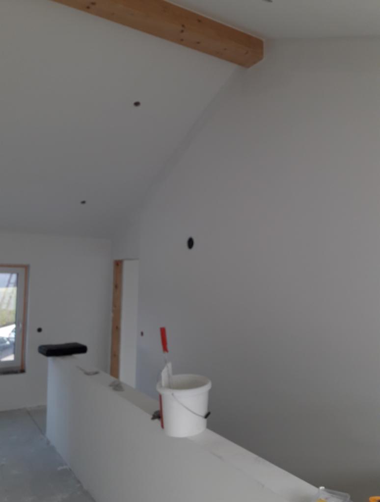 Wnętrze po malowaniu na biało, widoczny wiadro z farbą i wałek oparty o ścianę, surowa belka drewniana przy suficie.