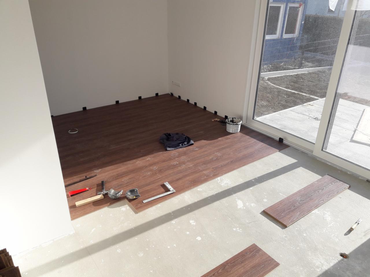 Proces układania paneli podłogowych w jasnym pomieszczeniu z widocznymi narzędziami: młotek, miarka kątowa, dobijak, oraz podkładki dystansowe przy ścianach.