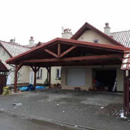 Drewniana pergola przy domu szeregowym z czerwonym dachem, widoczne przygotowane materiały budowlane i częściowo otwarty garaż.