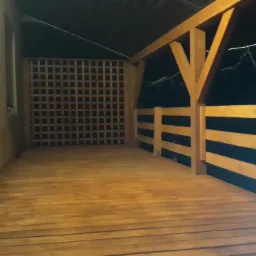 Zadaszona drewniana konstrukcja z kratownicą w tle, oświetlona ciepłym światłem, widok na nocne niebo.