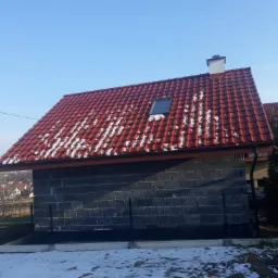 Niedokończony budynek z czerwoną dachówką, częściowo pokrytą śniegiem, widok na dach z oknem dachowym i kominem, ściany z szarych pustaków, czarne metalowe ogrodzenie i resztki śniegu na trawie.