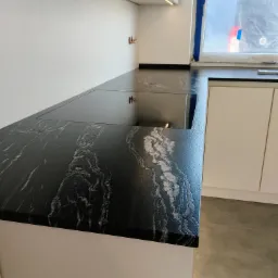 Czarny blat kamienny z białymi żyłkami, wbudowana płyta grzewcza, nowoczesna kuchnia, jasne szafki, okno z folią ochronną.