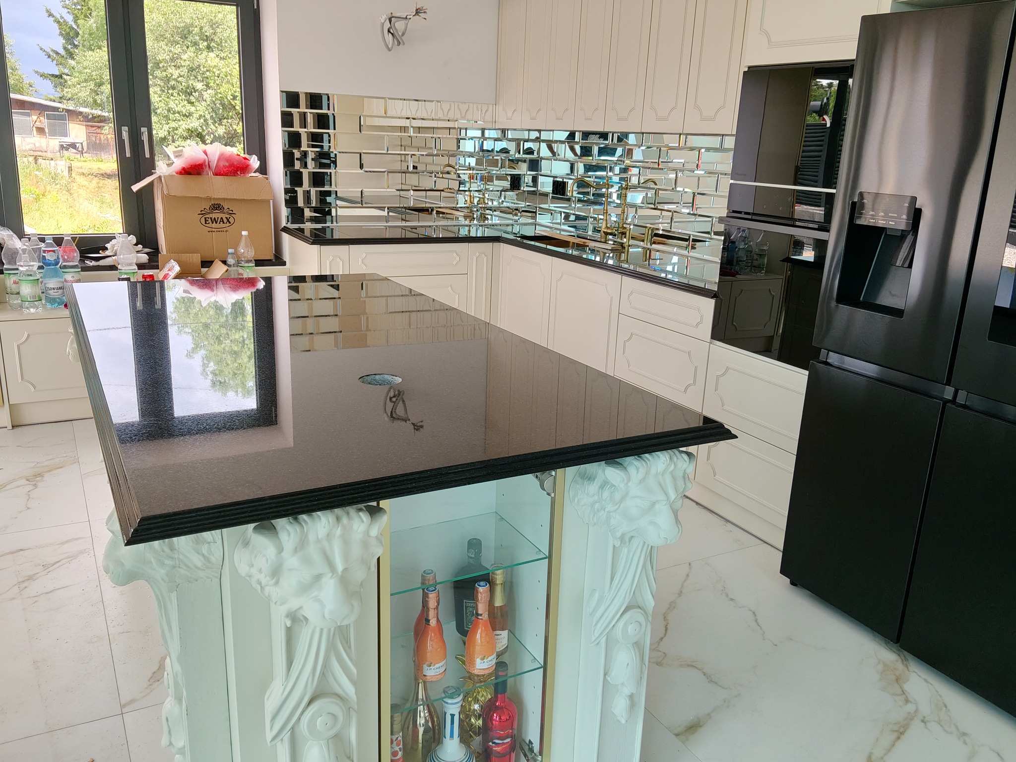 Nowoczesna kuchnia z czarnym blatem kamiennym na wyspie z dekoracyjnymi kolumnami i lustrzanymi płytkami na ścianie, uzupełniona czarną lodówką.