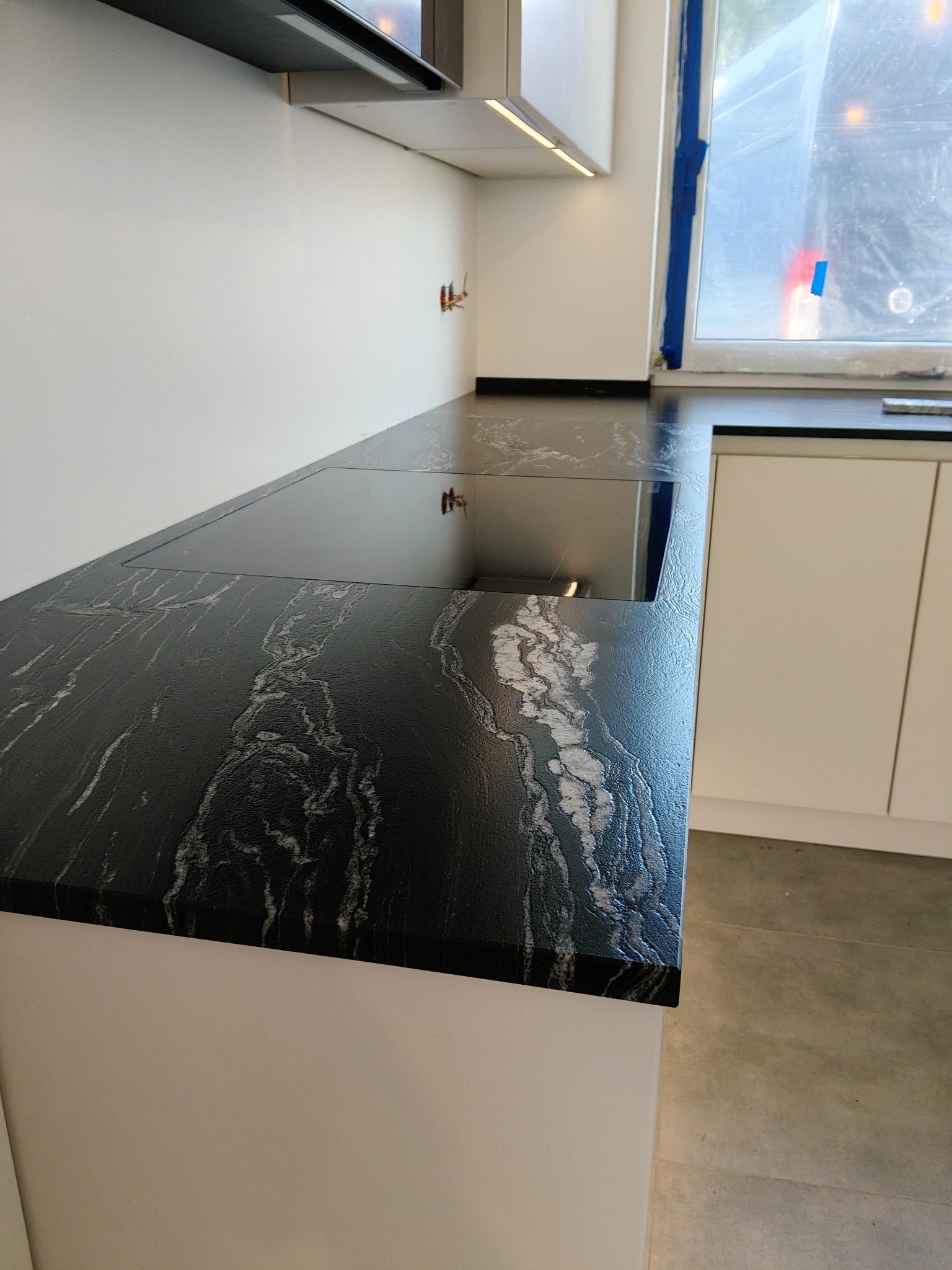 Czarny blat kamienny z białymi żyłkami, wbudowana płyta grzewcza, nowoczesna kuchnia, jasne szafki, okno z folią ochronną.