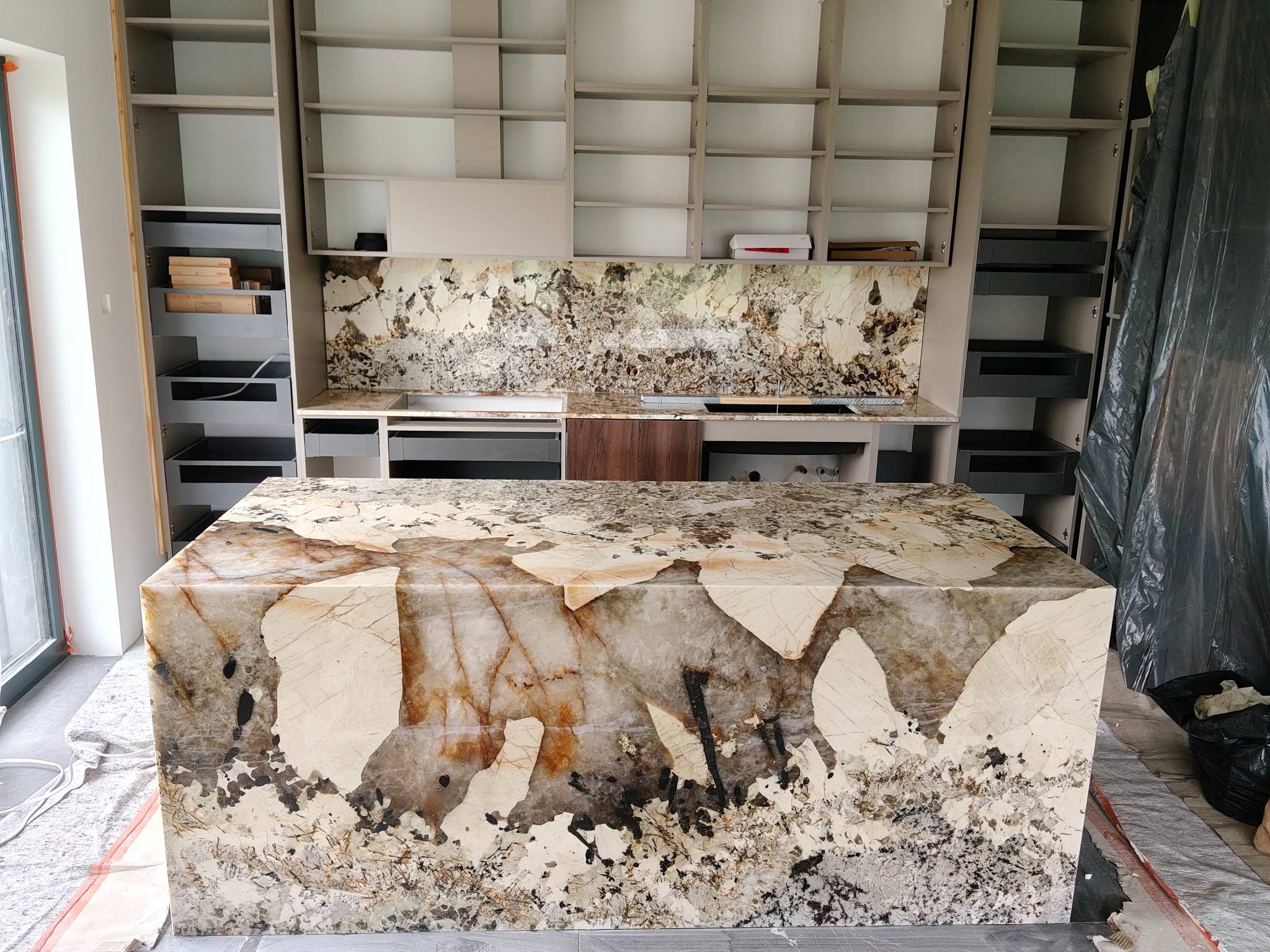 Blat kamienny z unikalnym wzorem, częściowo zamontowany w kuchni z szarymi szafkami. W tle widoczna ściana z kamiennym panelem o podobnym wzorze.