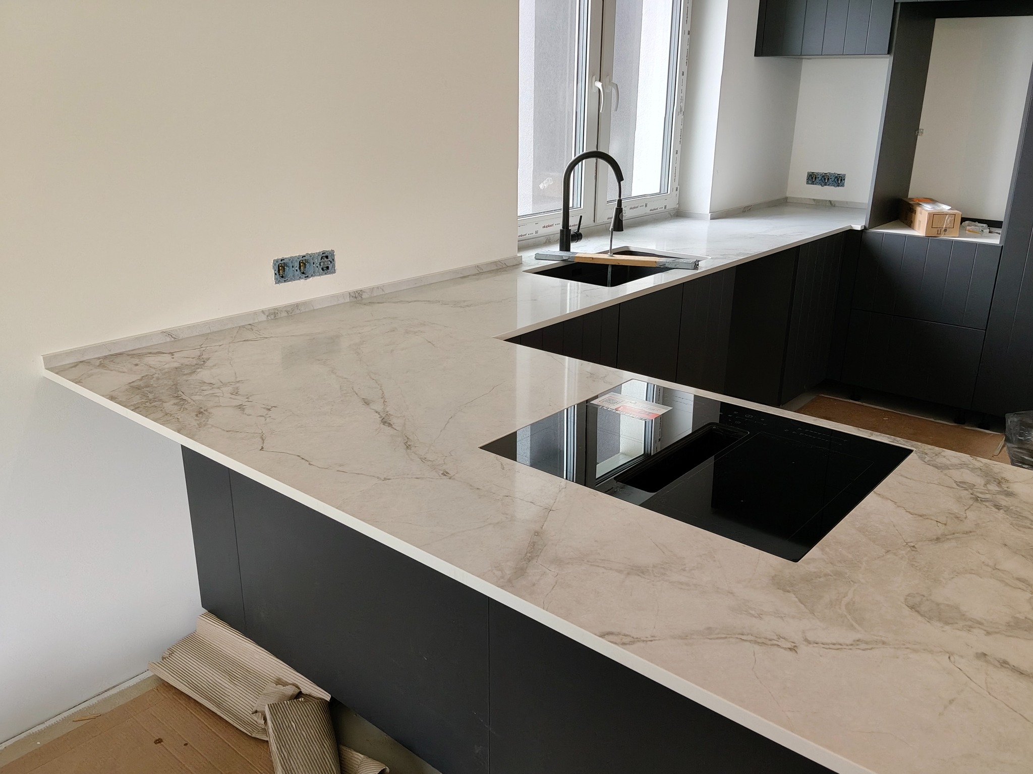 Jasny blat kuchenny z marmurowym wzorem, wbudowany zlew i płyta indukcyjna, czarne szafki, surowe ściany z instalacją elektryczną, nowoczesny design.