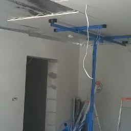 Montaż płyty gipsowo-kartonowej na suficie za pomocą podnośnika, widoczne profile metalowe, izolacja termiczna i fragment ściany z otworem drzwiowym.