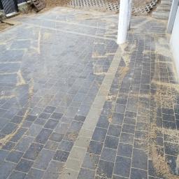 Przedsiębiorstwo usługowe - Ułożona, szara kostka brukowa z jasnoszarym pasem pośrodku, zasypana piaskiem, z widocznym słupem i fragmentem budynku w tle.