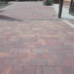 Ułożona nawierzchnia z kostki brukowej w odcieniach czerwieni i brązu, z widocznym odpływem liniowym i fragmentem ogrodu w tle.