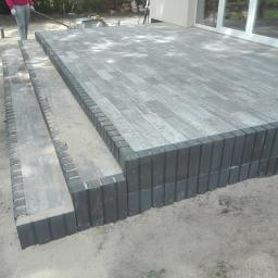 Przedsiębiorstwo usługowe - Wykończony taras z szarej kostki brukowej z obrzeżem z ciemniejszych elementów, widoczne schody z kostki, piasek wokół, częściowo widoczna osoba z łopatą w tle.