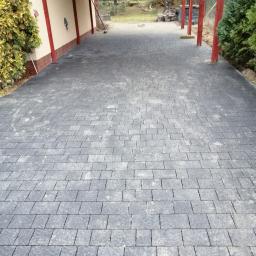 Przedsiębiorstwo usługowe - Ułożona szara kostka brukowa na podjeździe, widoczne czerwone filary zadaszenia i fragment budynku z zielonymi krzewami.