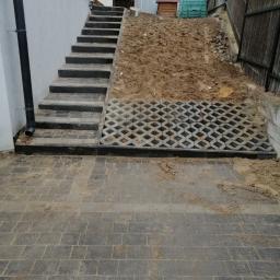 Przedsiębiorstwo usługowe - Układanie szarej kostki brukowej na podjeździe, widoczne schody z kostki prowadzące w górę, po prawej stronie ziemia i fragment ażurowej kostki brukowej.