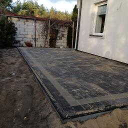 Przedsiębiorstwo usługowe - Niedokończona nawierzchnia z czarno-szarej kostki brukowej ułożona przy białym budynku, widoczna warstwa piasku na kostce i wykopana ziemia wokół.