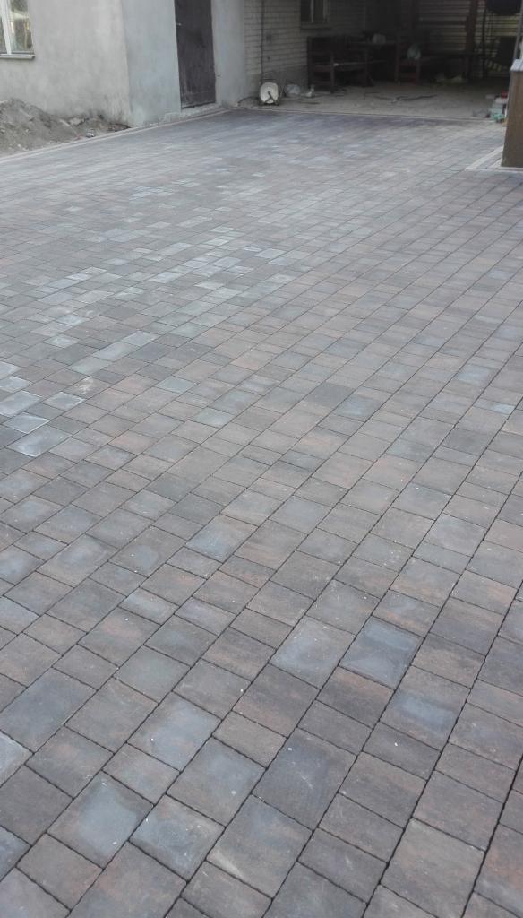 Nowo ułożona, brązowo-szara kostka brukowa na podjeździe, z widocznym budynkiem w tle i delikatnym oświetleniem.