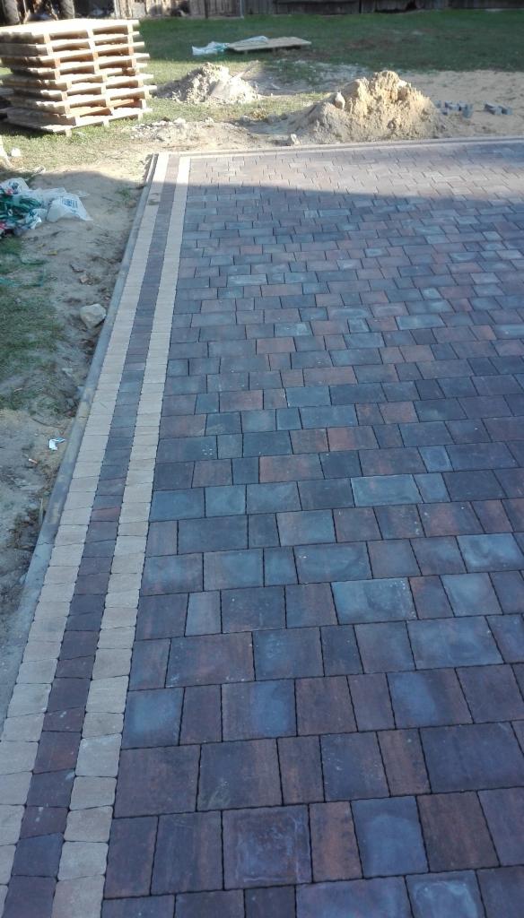 Ułożona, wielobarwna kostka brukowa z obrzeżem z jasnych kostek, widoczny fragment trawnika, paleta i pryzma piasku w tle.