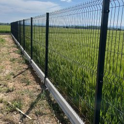 Lukas - Długi, czarny, metalowy płot panelowy z podmurówką z prefabrykatów betonowych na skraju pola uprawnego w słoneczny dzień.